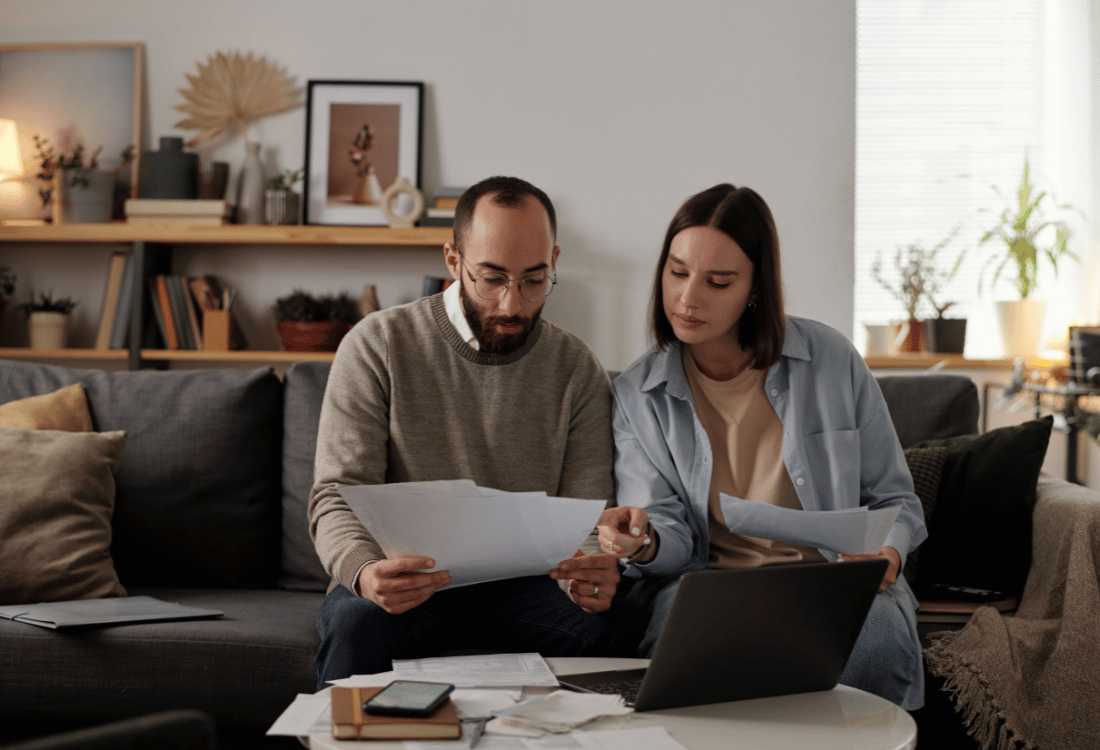 A man and woman sit together on a sofa, looking at paperwork and a laptop, reviewing council tax documents at home.