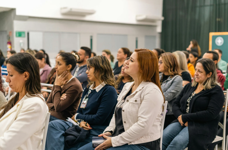 Residents seated in rows at a community meeting, listening attentively to a speaker out of frame in a bright indoor hall.