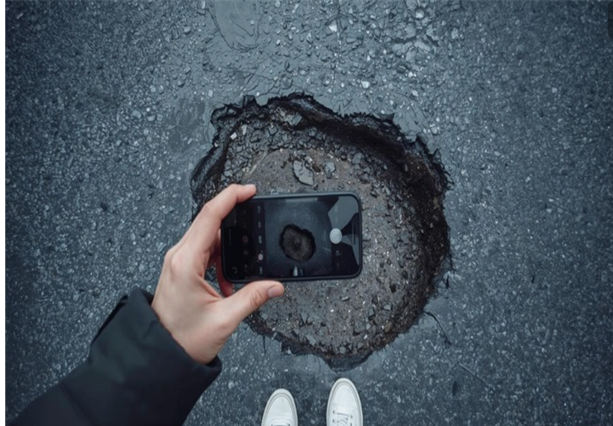 A person takes a photo of a large pothole on the road using a smartphone while wearing white trainers .