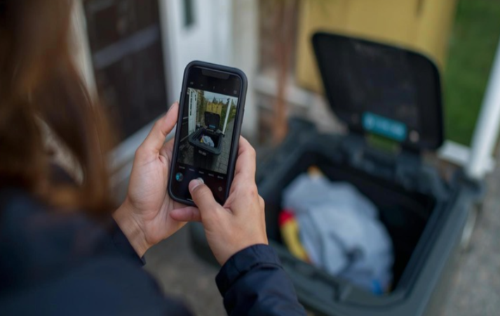 Person taking a photo of an open wheelie bin filled with rubbish outside a house.