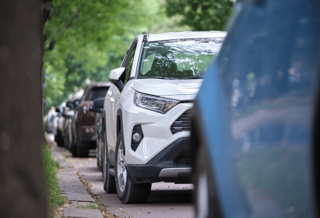 White SUV parked closely between other cars along a leafy residential street.